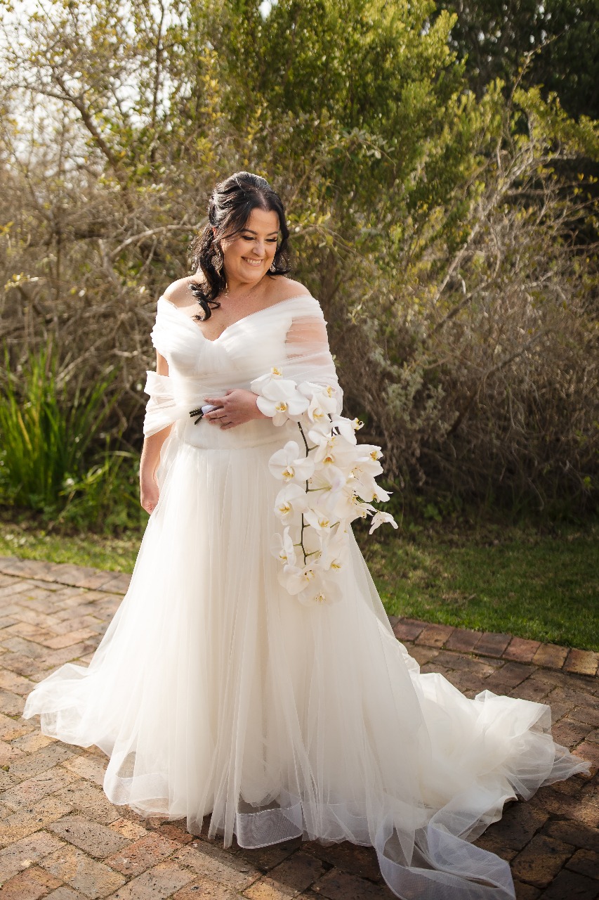 kim Ruched Tulle A-Line Gown with Off-the-Shoulder Elegance The Kim gown is a show-stopping statement of soft structure and modern romance. Designed with layers of shimmer tulle and delicate ruching, this off-the-shoulder beauty features a flattering drop waist and dreamy movement. The wide horsehair hemline adds volume and definition, making every step unforgettable. ✨ Design Highlights: ✔ Ruched Tulle Bodice – Sculpted to flatter with soft, elegant folds and a supportive fit. ✔ Off-the-Shoulder Straps – Romantic draped straps that highlight the collarbone and shoulders. ✔ Drop Waist Silhouette – Elongates the torso for a modern, flattering shape before flowing into a soft A-line. ✔ Shimmer Tulle Layers – A subtle sparkle throughout the skirt adds a radiant glow. ✔ Ruched Tulle Bolero – Optional matching ruched tulle bolero for added coverage or drama. ✔ Wide Horsehair Hemline – Creates structured movement and a crisp, couture finish to the skirt. ✔ Zip & Button Back Closure – Smooth fit with classic bridal button detailing. 💫 Customization Options: ✅ Custom fit to your exact measurements ✅ Option to remove bolero or adjust strap style ✅ Available in ivory, soft white, or blush shimmer tulle ✅ Train length and hemline width can be customized 💎 Kim is made for the bride who wants soft glamour, modern sophistication, and a dash of drama on her special day. 💎 📞 Book your fitting or custom consultation today! 📧 info@bestersbridal.co.za | 📱 084 268 1354 Ruched Tulle • Drop Waist • Off-Shoulder Straps • Shimmer Layers • Horsehair Hem • A-Line Silhouette