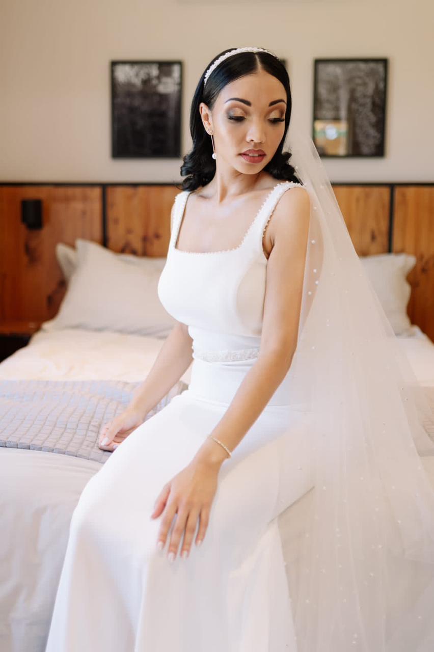 Crepe Sheath Gown with Pearl Trim and Elegant Square Neckline The Tercia gown is the epitome of understated luxury—sleek, modern, and timelessly graceful. Designed in soft stretch crepe, this figure-skimming sheath silhouette features refined pearl detailing for a bridal look that is both contemporary and classic. ✨ Design Highlights: ✔ Square Neckline Bodice – Clean and structured, offering a flattering frame to the collarbone and shoulders. ✔ Pearl Bead Embellishments – Delicate pearl beading decorates the neckline, straps, and back bodice for a subtle touch of bridal shimmer. ✔ Fitted Sheath Silhouette – Effortlessly elegant, gently contouring the body for a graceful and elongating look. ✔ Zipper Back Closure – Neatly finished with a sleek fit and invisible zip. ✔ Pearl-Beaded Bridal Belt – A matching belt enhances the waist and adds extra refinement. ✔ Small Sweep Train – A soft, graceful train ideal for minimalistic elegance without the weight. 💫 Customization Options: ✅ Available in ivory or soft white crepe ✅ Custom sizing for a perfect tailored fit ✅ Option to adjust neckline or train length ✅ Add a matching pearl-trimmed veil for a complete look 💍 Tercia is designed for the bride who values simplicity with elegance—where every detail, down to the last pearl, speaks of refined bridal beauty. 💍 📞 Book your exclusive fitting or design consultation today! 📧 info@bestersbridal.co.za | 📱 084 268 1354 Square Neckline • Pearl Beaded Trim • Sheath Silhouette • Bridal Belt • Small Train • Zipper Back